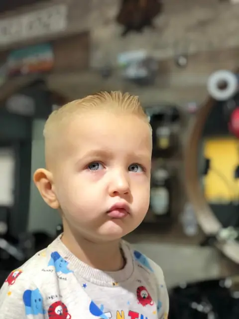 Băiețel blond cu tunsoare scurtă, privind spre cameră